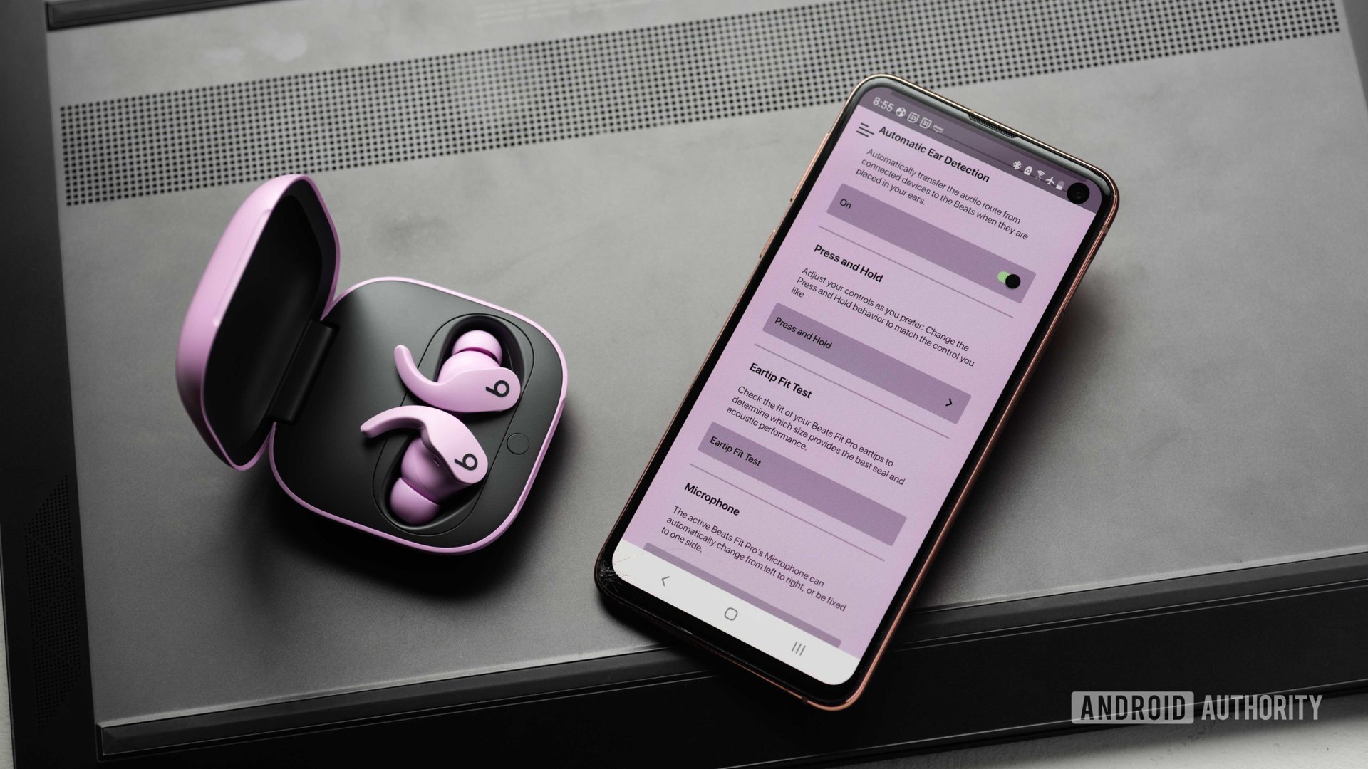 在开放的充电盒中取消真正的无线耳罩，旁边是三星Galaxy S10E，带有击败App打开的击败。该应用程序对其具有紫色色调，可能是匹配耳机。